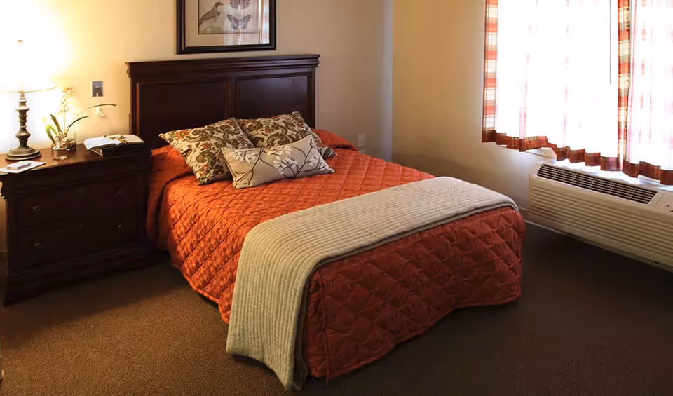 A cozy bedroom with a wooden headboard bed covered in an orange quilt and decorative pillows. There is a nightstand with a lamp and a small plant on it next to the bed. A window with plaid curtains and an air conditioning unit is on the right side of the room.