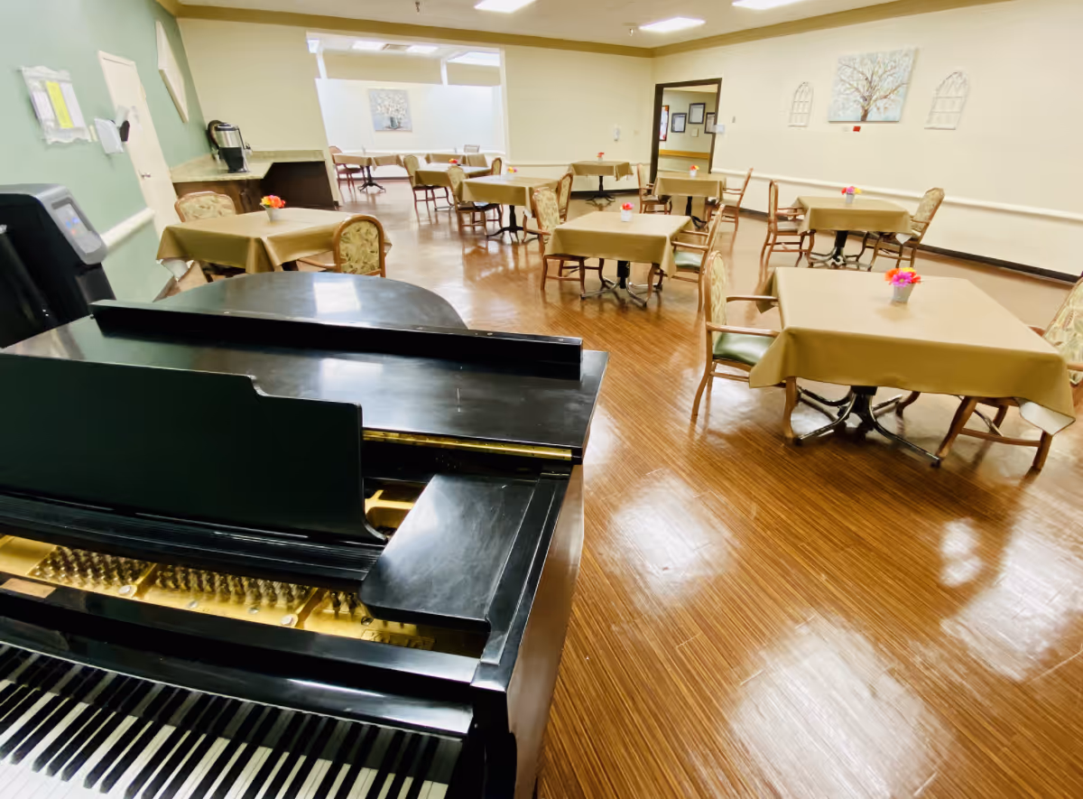 Interior dining/activity room with a black grand piano in the foreground and multiple small tables with beige tablecloths and chairs on a polished wood floor.