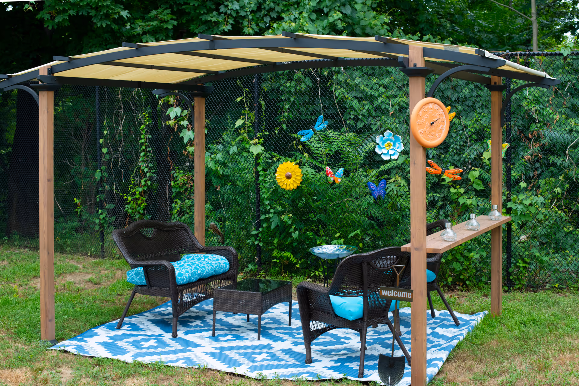 Outdoor seating area with a wooden pergola covered by a beige canopy. Underneath, there are two black wicker chairs and a loveseat with blue patterned cushions, a small black wicker coffee table, and a blue and white patterned outdoor rug. The pergola has a wooden shelf with four glass candle holders and a round orange thermometer attached to one of the wooden posts. Decorative metal butterflies and flowers are attached to a black chain-link fence covered with green foliage in the background. A small shovel with a 'welcome' sign is placed near one of the chairs.