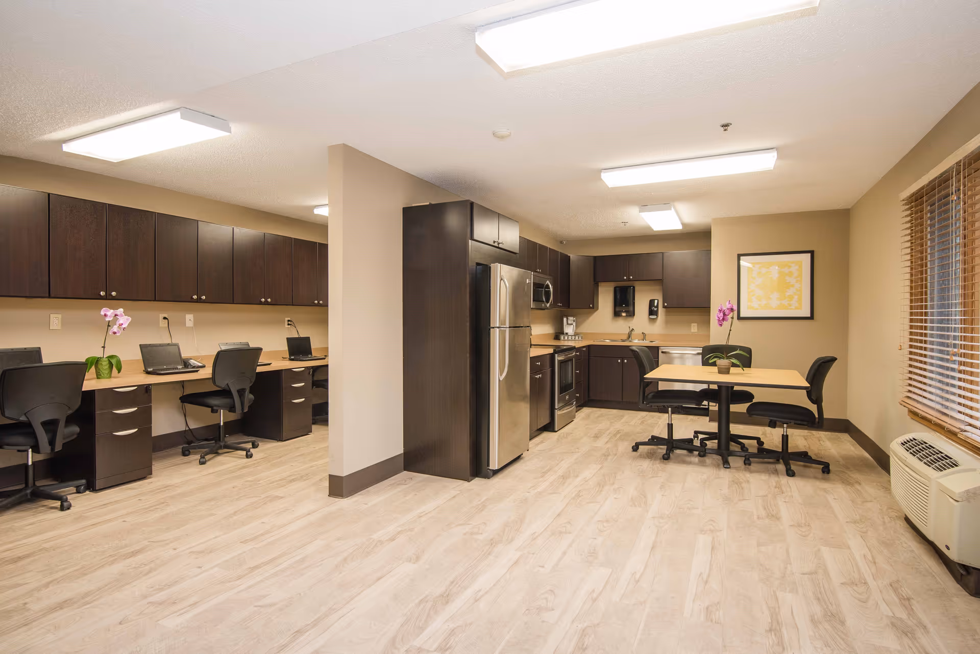 A spacious room featuring a kitchen area with stainless steel appliances including a refrigerator, stove, microwave, and dishwasher. Adjacent to the kitchen is a small table with two chairs and a potted orchid. On the left side of the room, there is a long desk with multiple office chairs and laptops, with dark wood cabinets above and below the desk. The room has light wood flooring, beige walls, and a window with wooden blinds on the right side.