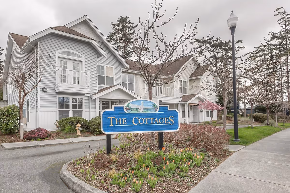 Two-story cottage-style building with a blue sign reading "The Cottages" in landscaped grounds beside a sidewalk and lamppost.