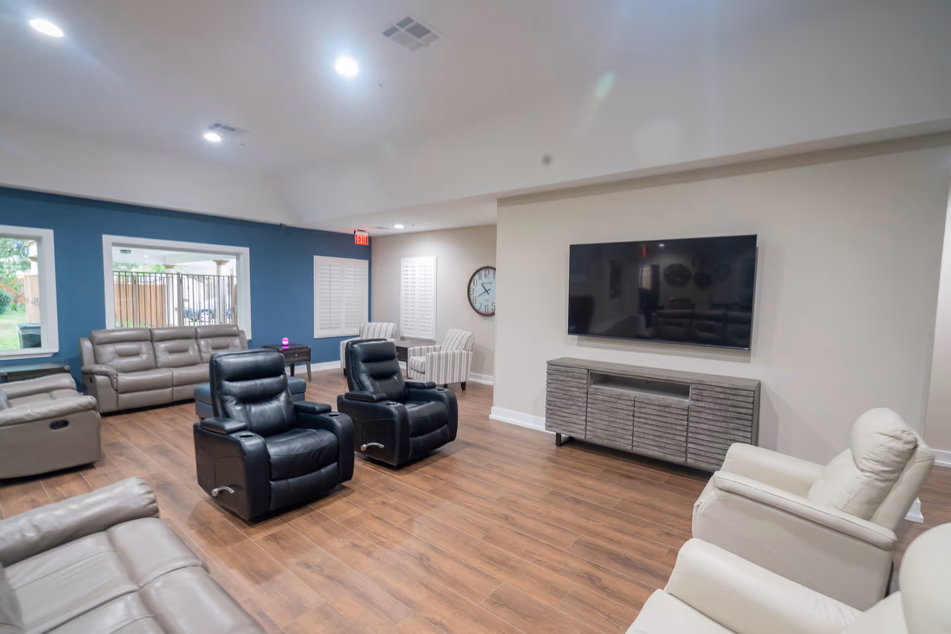A spacious living room with multiple recliner chairs and sofas arranged around a large flat-screen TV mounted on the wall. The room has wooden flooring, a blue accent wall with windows, and a large clock on a white wall. The space is well-lit with recessed ceiling lights.