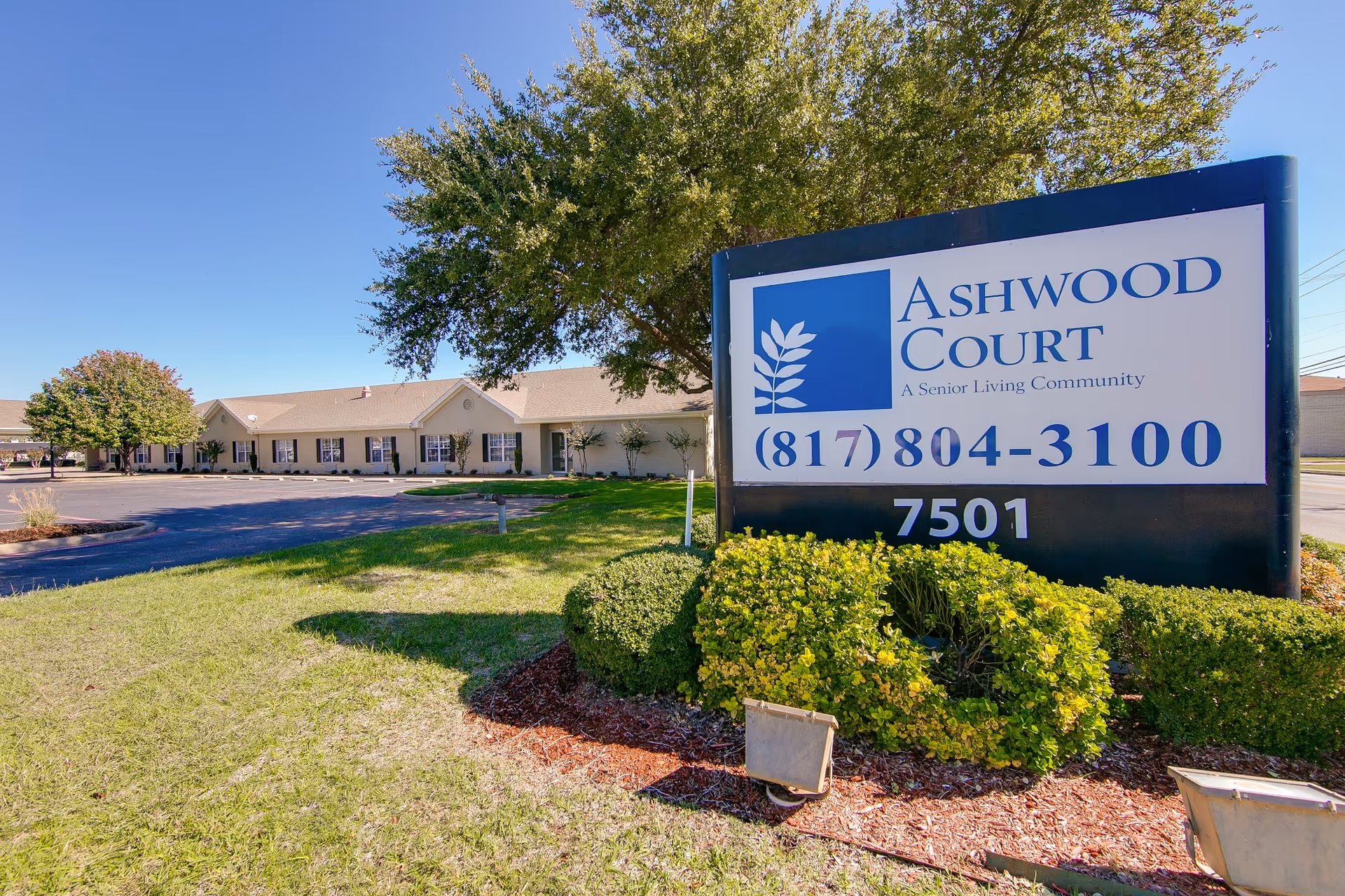 Outdoor view of Ashwood Court senior living community sign with phone number and address, surrounded by green bushes and grass, with the facility building and clear blue sky in the background.