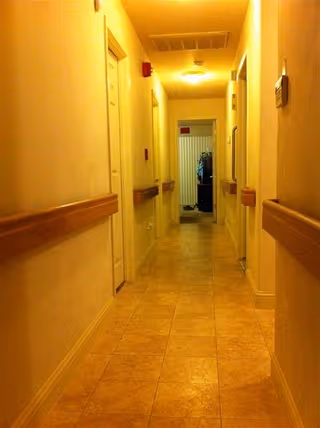 A narrow indoor hallway with beige tiled floor and cream-colored walls. There are wooden handrails on both sides of the hallway, several closed doors along the walls, and a ceiling light fixture. At the end of the hallway, there is an open door revealing another room with some items visible inside.