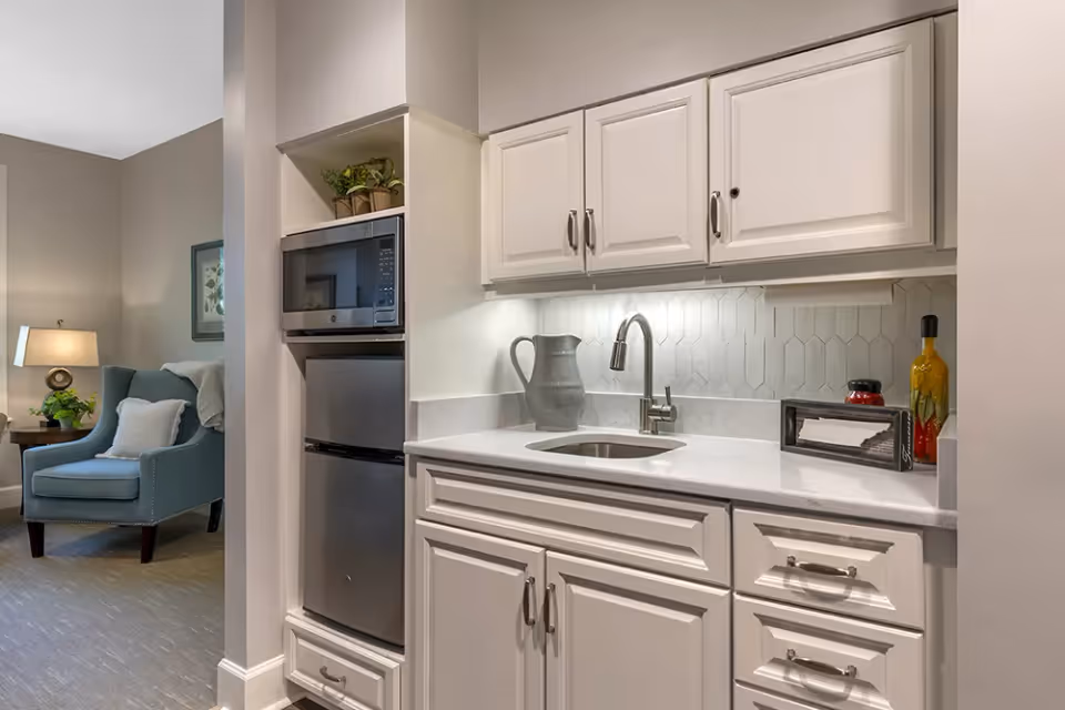 A small kitchenette area with white cabinets, a stainless steel mini refrigerator, and a microwave. The countertop has a sink with a modern faucet, a gray pitcher, and decorative items including a bottle and a small framed picture. In the background, there is a cozy living area with a blue armchair, a side table with a lamp, and a plant.