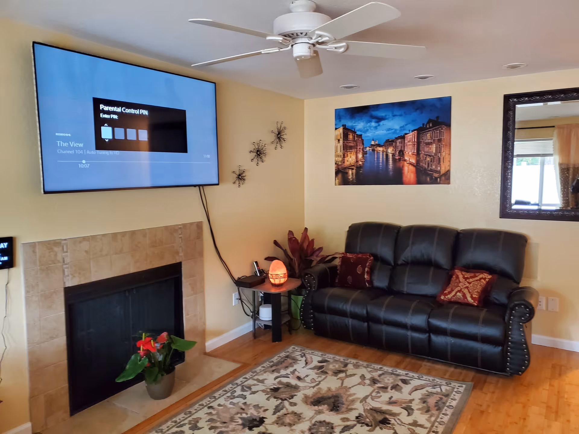 A cozy living room with a black leather couch adorned with two decorative pillows, a wall-mounted flat-screen TV displaying a parental control PIN screen, a fireplace with a potted plant on the hearth, a small side table with a glowing salt lamp and a plant, a ceiling fan, a large mirror on the wall, and a framed picture of a canal scene at dusk. The room has wooden flooring and a patterned area rug.