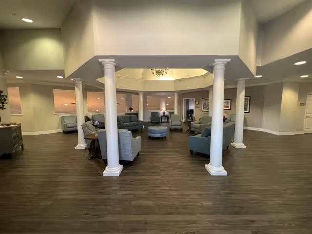 Open common lounge area with sofas, armchairs, ottomans and white columns under a high ceiling.