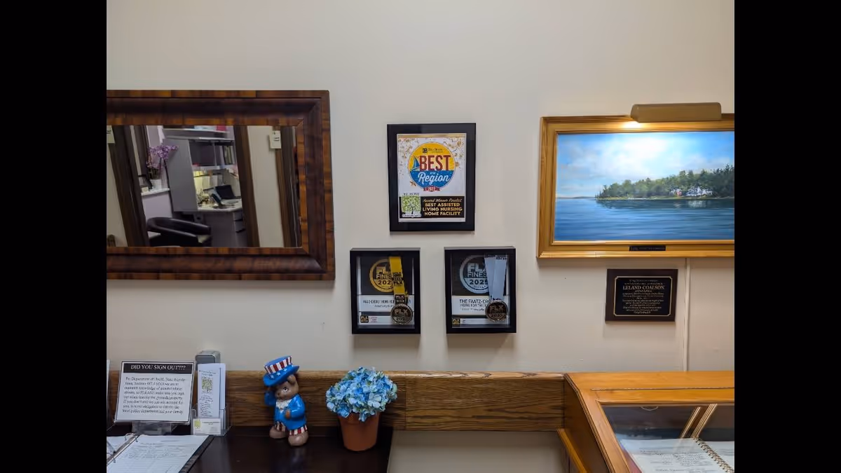 Interior wall of a senior living facility with a wooden-framed mirror reflecting part of an office area, three framed awards hanging on the wall, a framed painting of a coastal scene with a gold frame and light above it, a small plaque beneath the painting, a small figurine of a bear dressed in patriotic clothing, a potted blue hydrangea, and a sign-in clipboard on a wooden counter.
