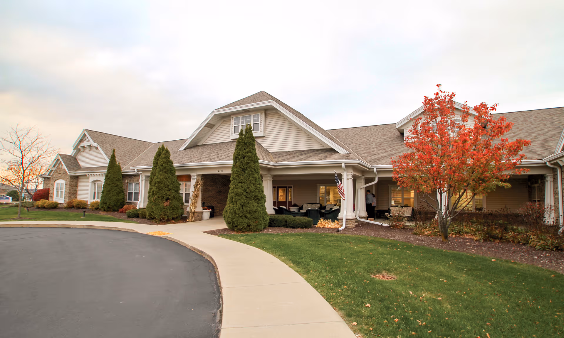 Exterior view of a single-story senior living facility building with a covered entrance, surrounded by green grass, trees including one with red autumn leaves, and a paved driveway and sidewalk.