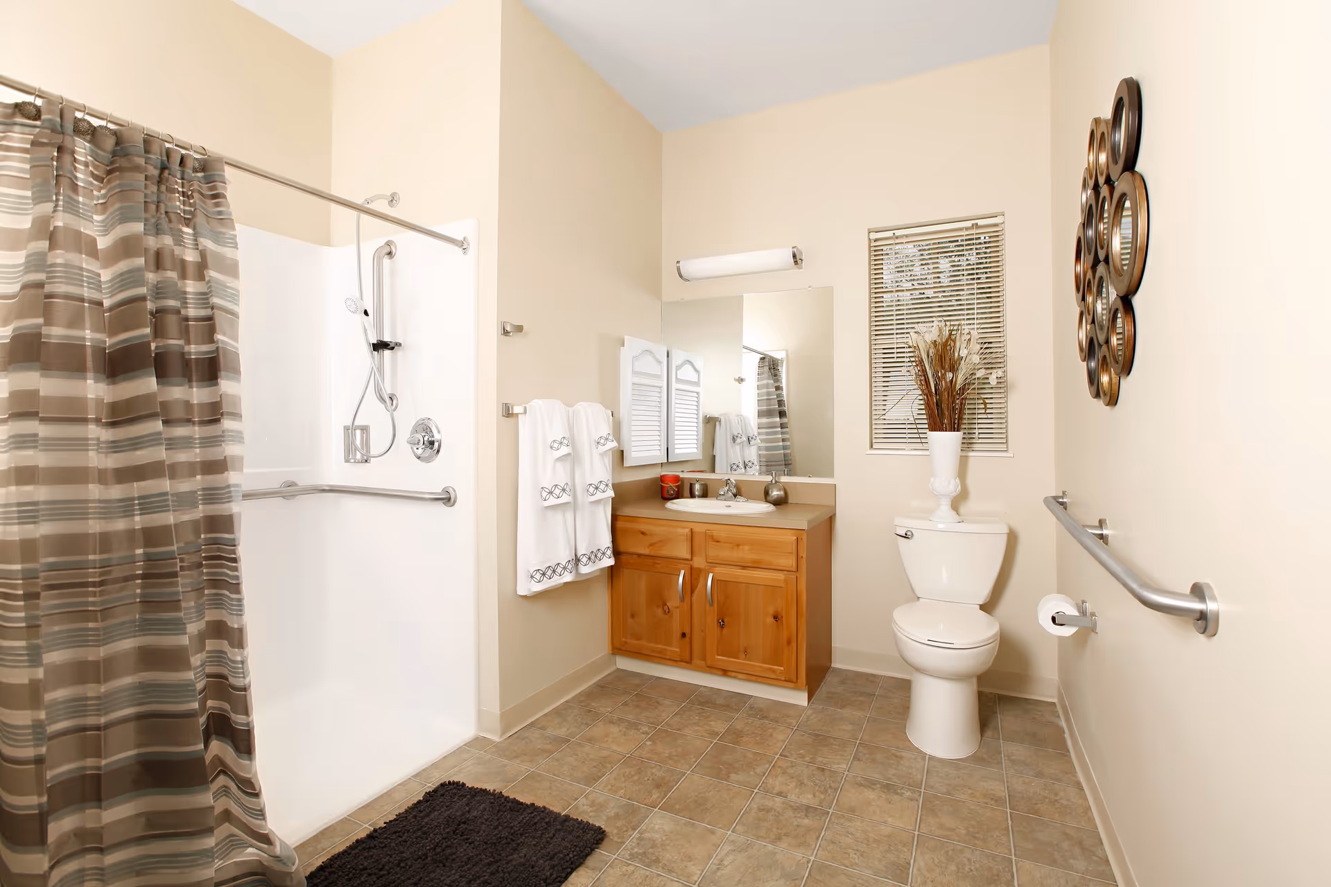 A clean and spacious bathroom with beige walls and tiled floor. It features a walk-in shower with a striped shower curtain, a wooden vanity with a sink and mirror above it, a toilet with a decorative vase on top, and a window with blinds. There are towels hanging on a rack and a grab bar next to the toilet for accessibility.