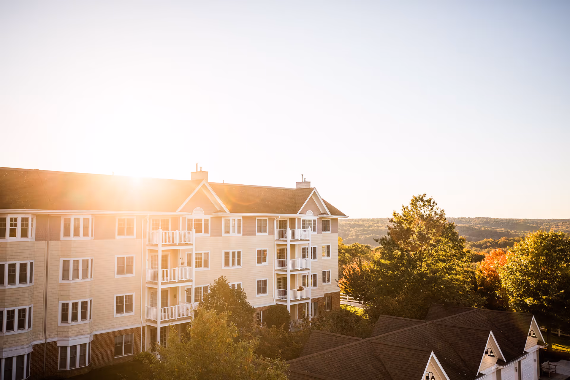 Exterior view of a multi-story residential building with balconies, surrounded by trees and greenery under a clear sky with the sun shining brightly.