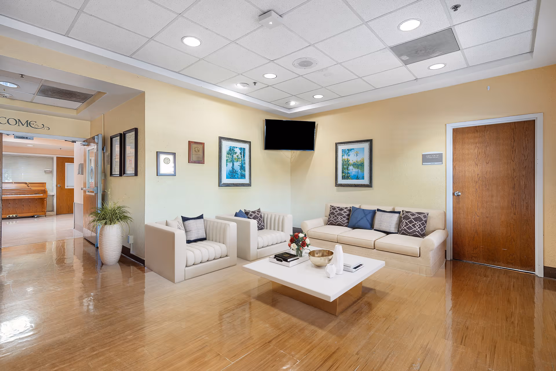 A cozy seating area in a senior living facility with two cream-colored armchairs and a matching sofa adorned with decorative pillows. A white coffee table with books, a bowl, and a flower arrangement sits in the center. The walls are light yellow with framed artwork and plaques. A flat-screen TV is mounted on the corner wall above the seating. The floor is polished wood, and there is a wooden door on the right side. To the left, a glass door leads to another room with a piano visible inside.