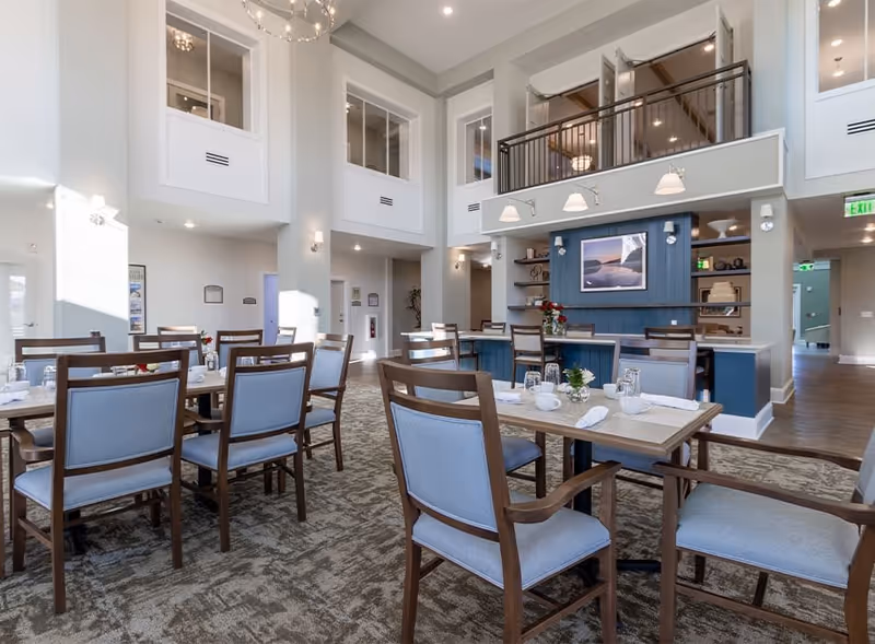Bright two-story dining area with multiple set tables, upholstered chairs, and a service counter beneath an interior balcony.