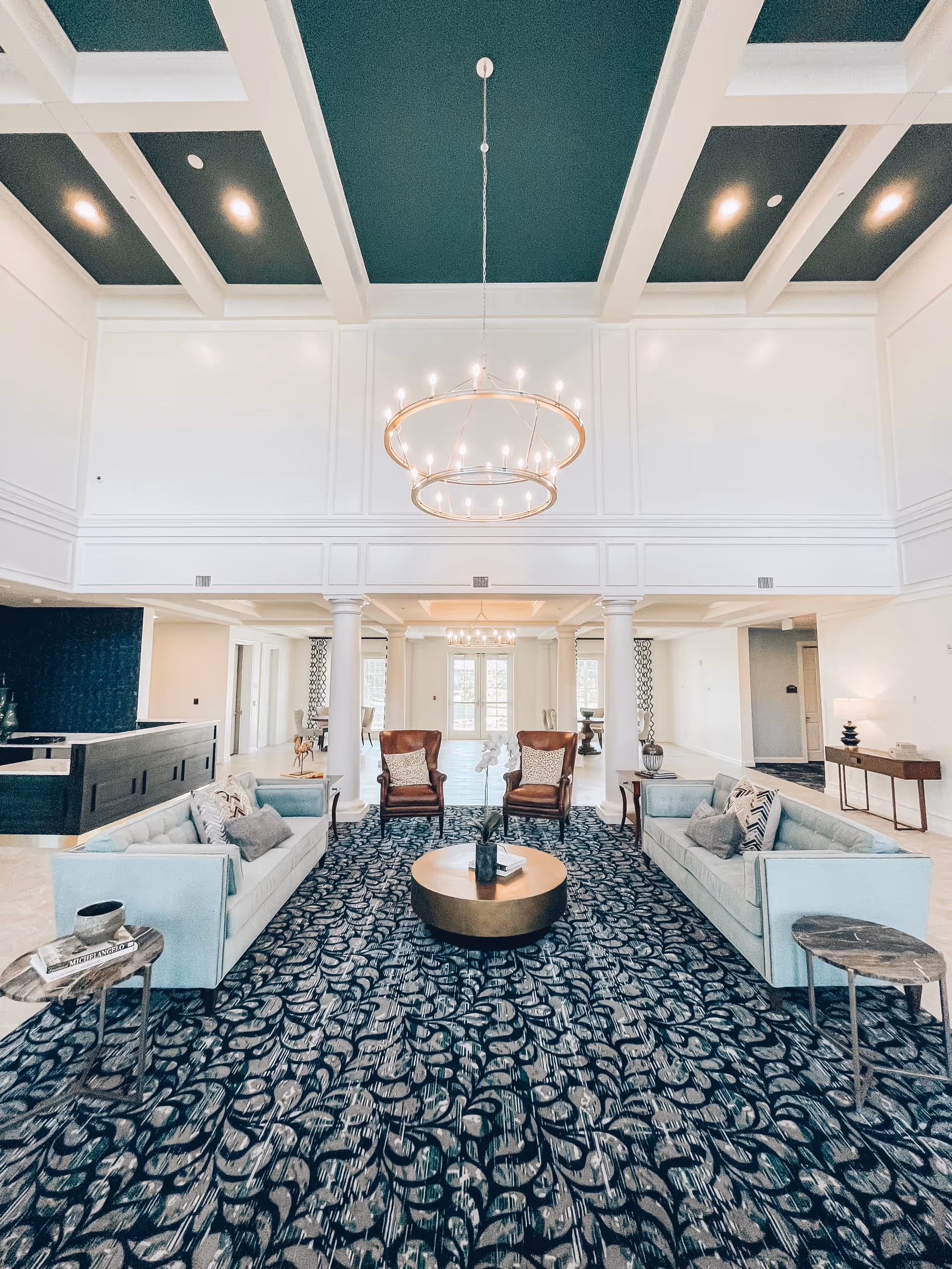 Spacious and elegant living room area with two light blue sofas facing each other, two brown leather armchairs, a round gold coffee table in the center, and a patterned dark carpet. The room features white walls, tall ceilings with green recessed panels, and two modern circular chandeliers hanging from the ceiling. There are white columns and additional seating visible in the background.