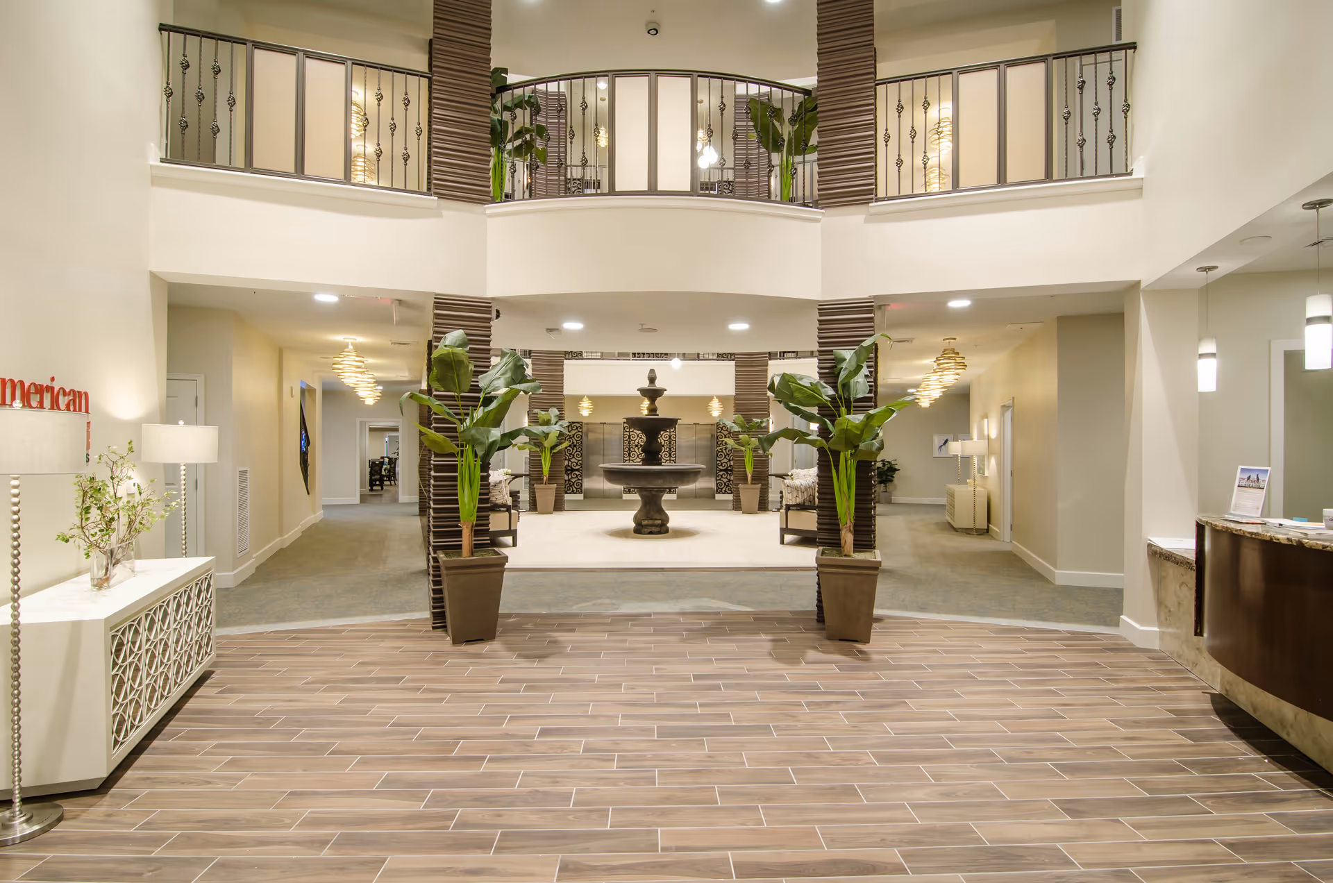 Spacious two-story senior living lobby with a central fountain, potted plants, seating areas, and a reception desk.