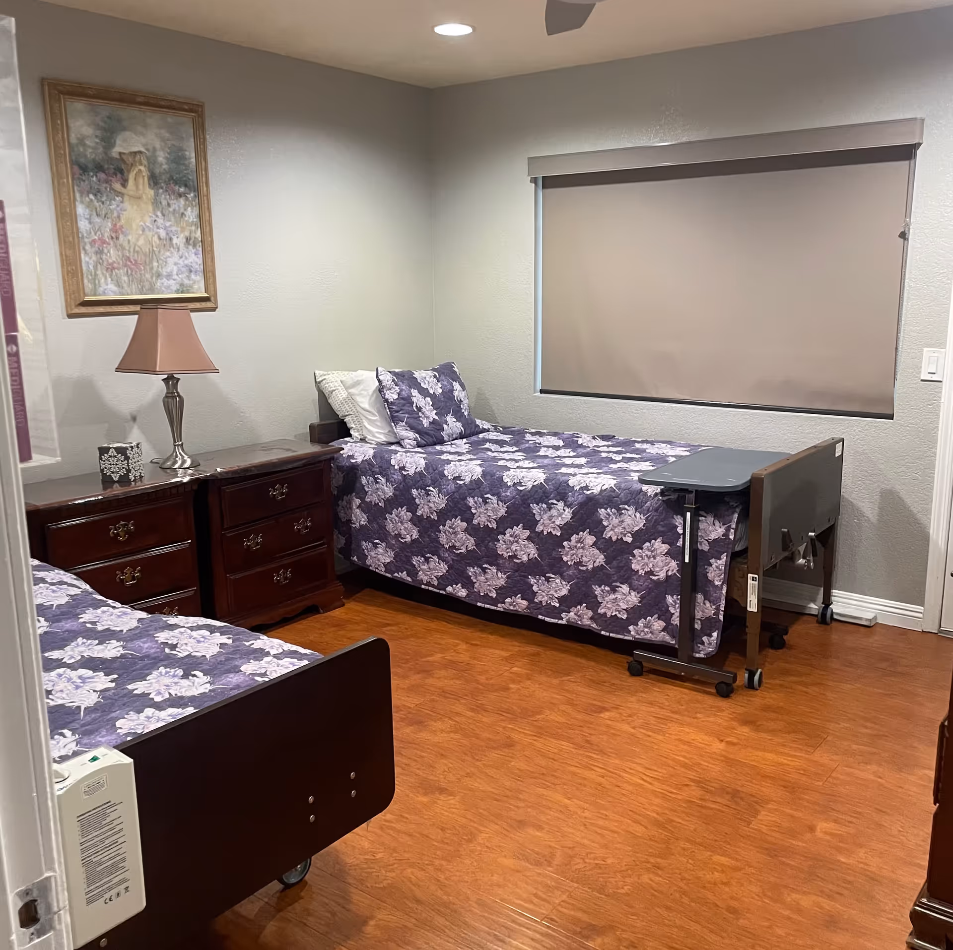 A bedroom with two twin beds dressed in purple floral quilts, a wooden nightstand with a lamp, a framed painting, and a closed window shade.