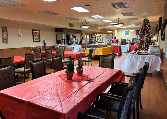 Dining room with multiple tables covered in red tablecloths, chairs, a buffet serving area in the back, and a decorated Christmas tree.