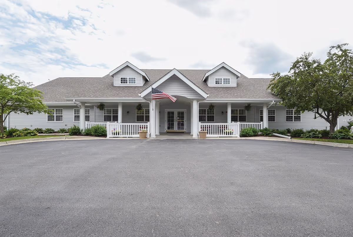 Front exterior view of a single-story senior living facility building with a gray roof, white siding, a covered entrance with white columns, an American flag, and landscaped greenery on both sides.
