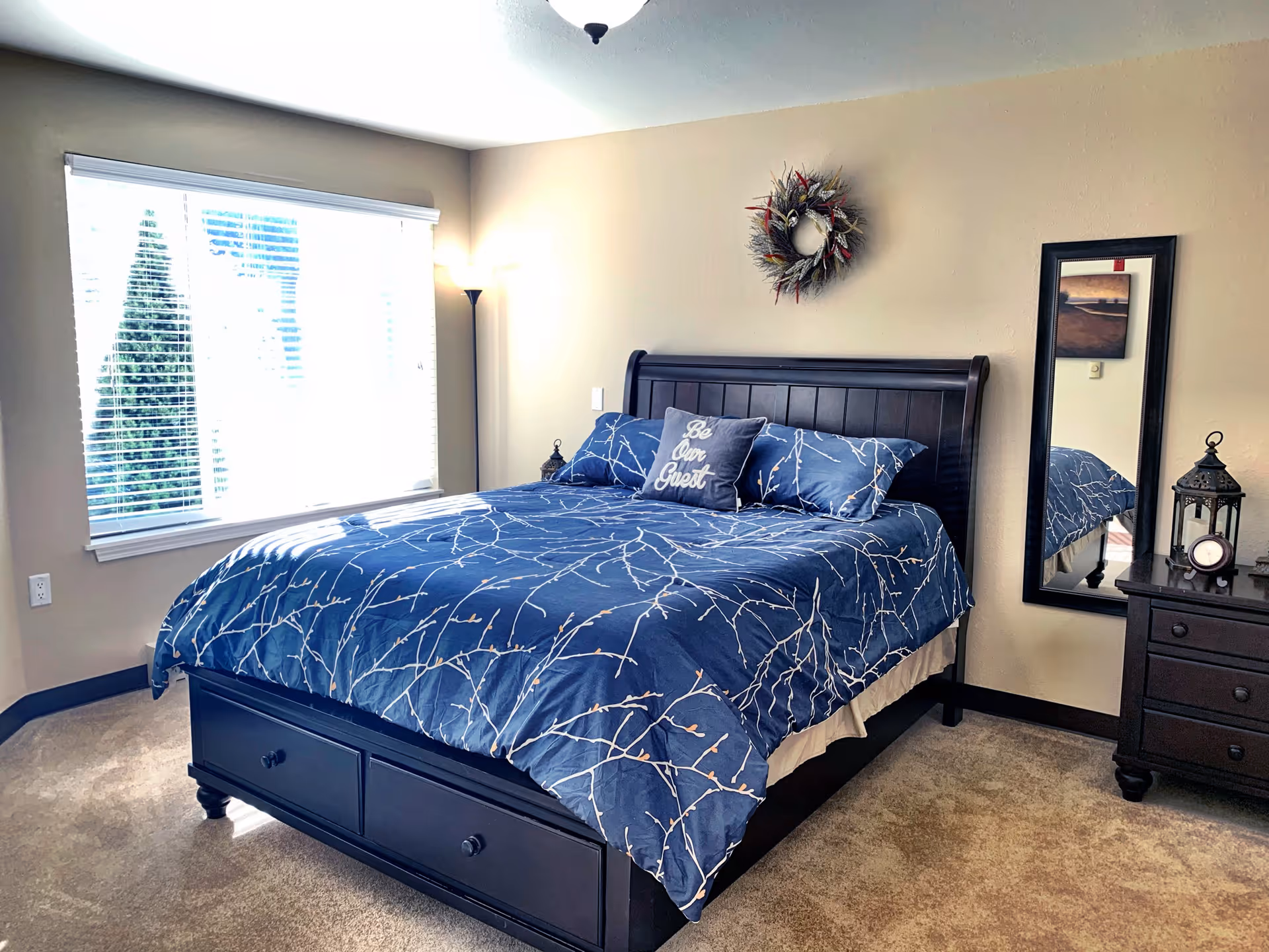 A bedroom with a large bed featuring a dark wooden frame and blue bedding with a white branch pattern. There are three pillows on the bed, one with the words 'Be Our Guest.' A window with blinds lets in natural light on the left side. A floor lamp stands in the corner, and a decorative wreath hangs on the beige wall above the bed. To the right, there is a dark wooden nightstand with a lantern and a clock, and a tall mirror is mounted on the wall.