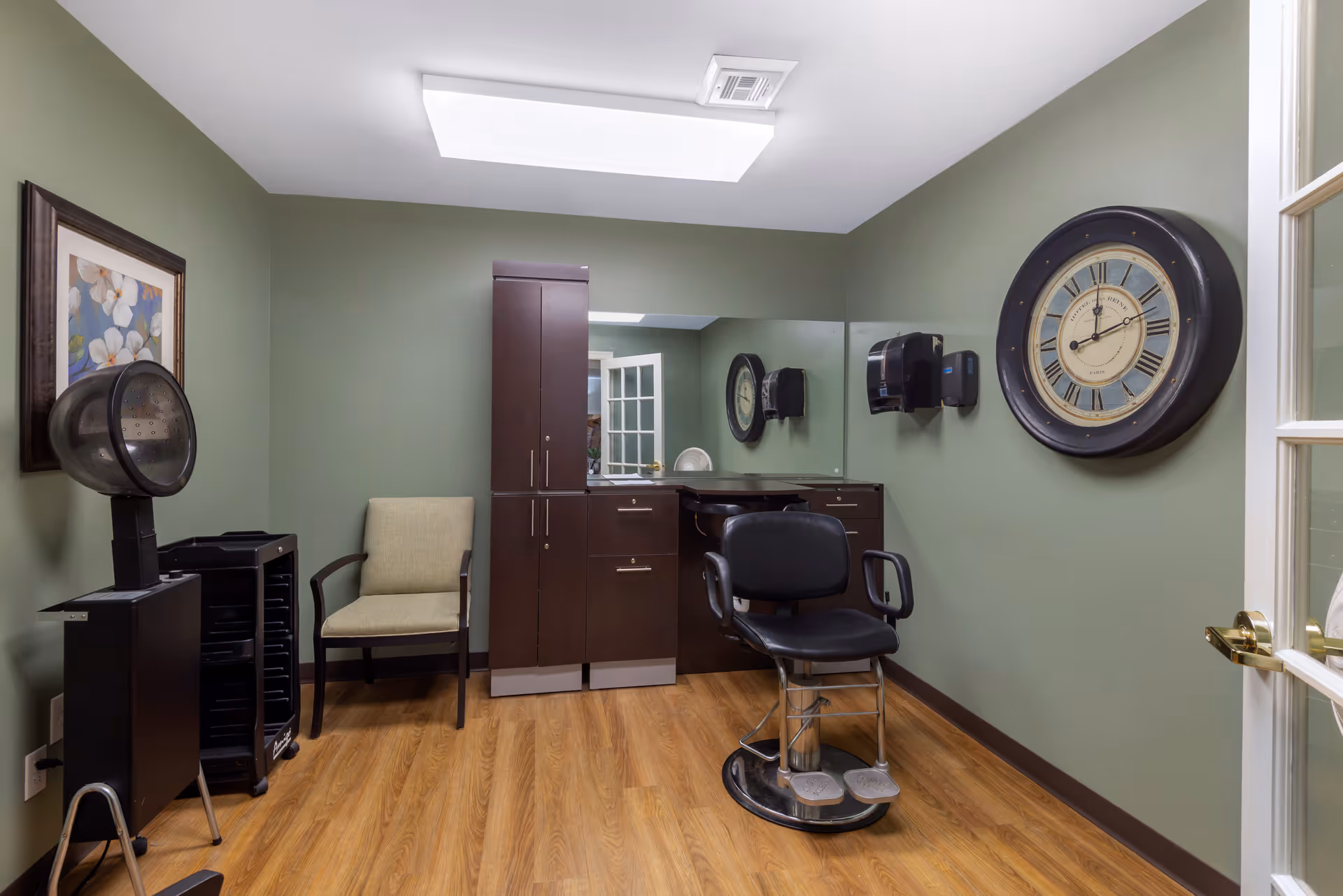 A small salon room with green walls and wood flooring. The room contains a black salon chair, a beige armchair, a hair dryer on a stand, dark wood cabinets with a large mirror above them, a large round wall clock, a wall-mounted paper towel dispenser, and a framed floral painting.
