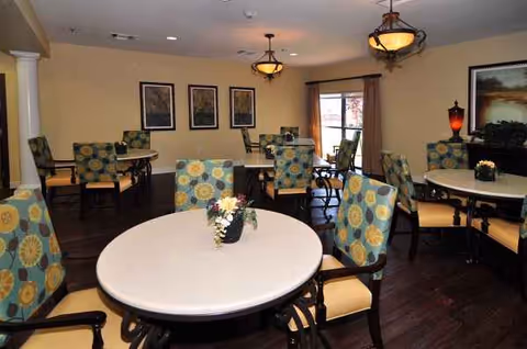 Interior dining room in a senior facility with round tables, patterned chairs, and centerpieces.