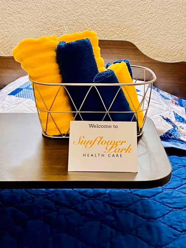 A wire basket containing neatly folded yellow and navy blue towels placed on a wooden tray on a bed with a blue quilt. In front of the basket is a card that reads 'Welcome to Sunflower Park Health Care'.