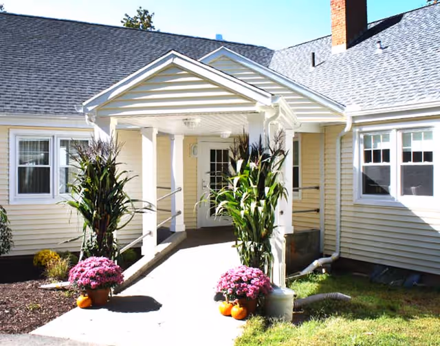 Entrance of a residential care facility with a covered porch supported by white columns. There are potted purple flowers and small pumpkins on either side of the walkway leading to a white door. The building exterior is light yellow with white trim and multiple windows.