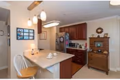 Interior view of a kitchen area with a white island countertop featuring two teacups and a lamp. The kitchen has dark wooden cabinets, a stainless steel refrigerator, and various kitchen appliances. There is a wooden chair at the island and a wooden dresser with decorative items on the right side. The lighting includes hanging pendant lights above the island and a ceiling light fixture.