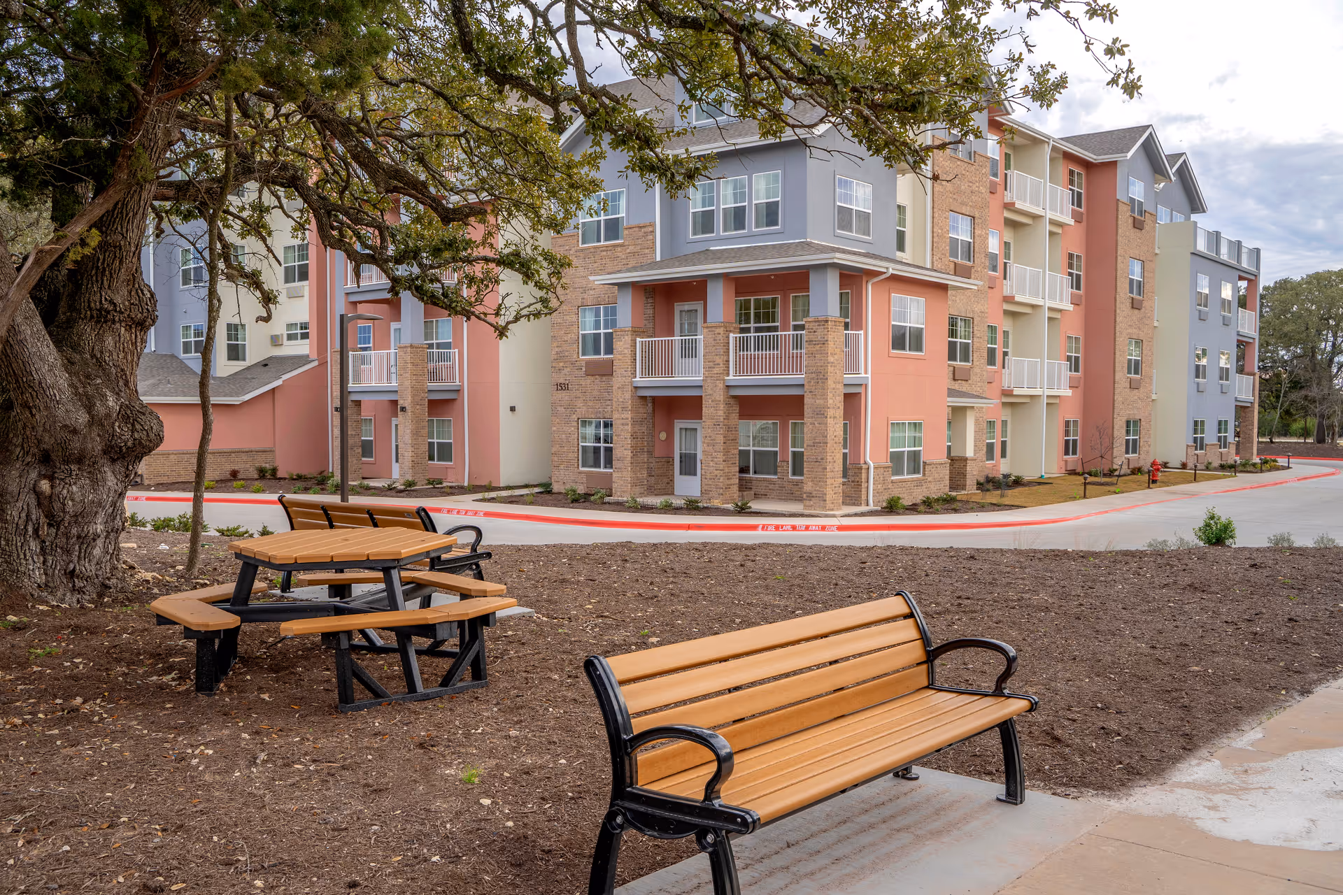 Outdoor seating area with wooden benches and picnic tables in front of a multi-story pastel-colored senior living building.