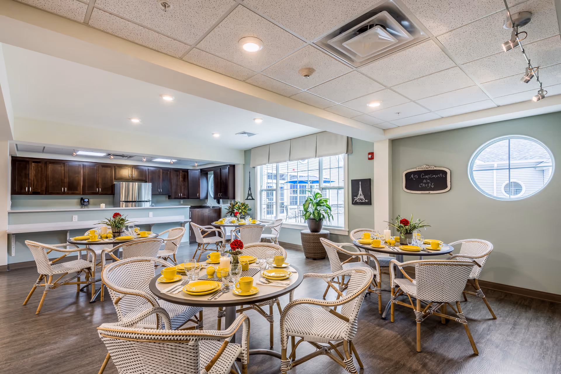 A bright dining room in a senior living facility with round tables set with yellow plates, cups, and silverware. White wicker chairs surround the tables. The room features a large window with a view of an outdoor patio, a kitchen area with dark wooden cabinets and stainless steel appliances, and light green walls with decorative wall art including an Eiffel Tower picture and a chalkboard sign.