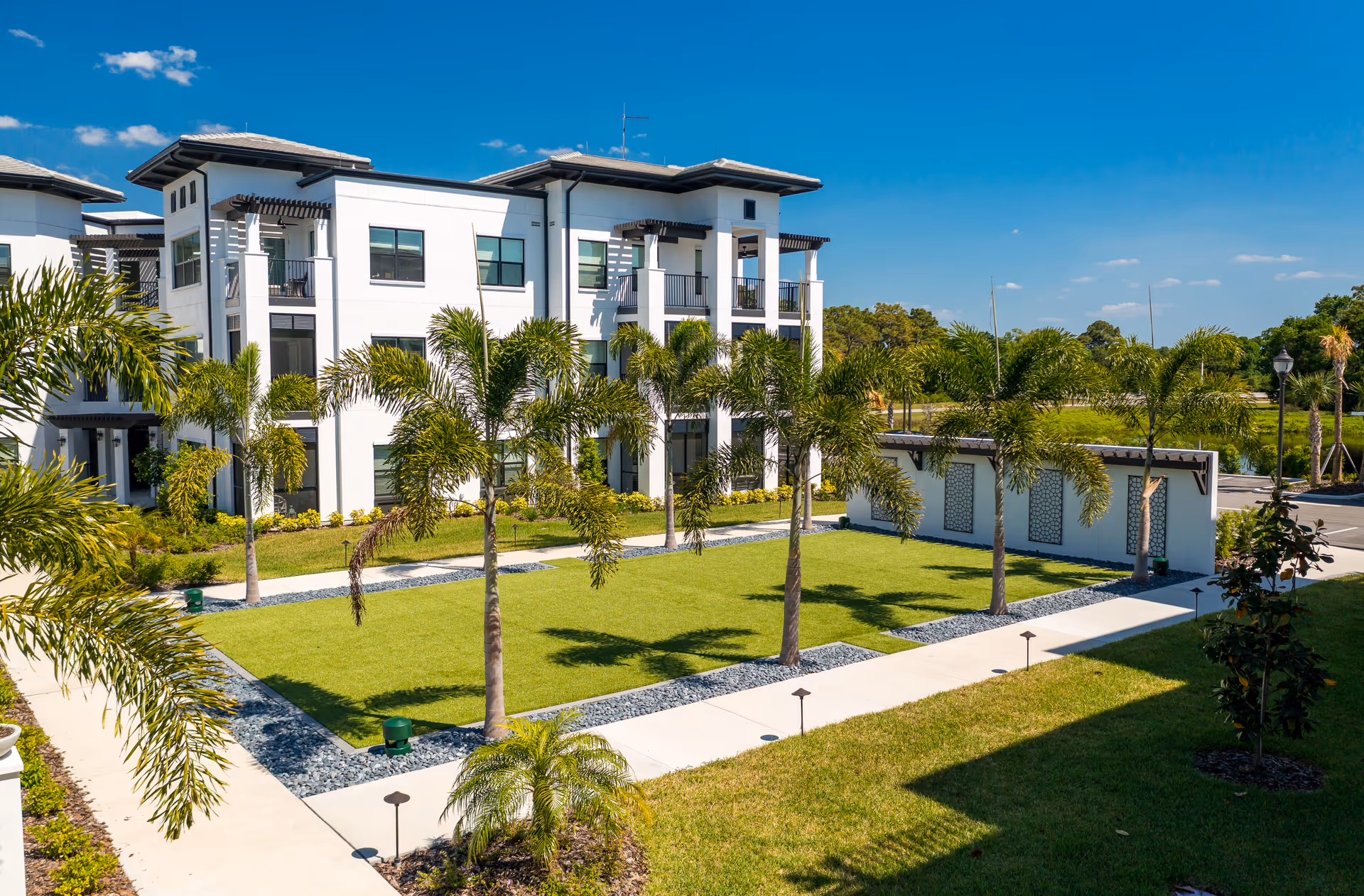 White three-story building with balconies overlooking a landscaped courtyard of palm trees and walkways under a clear blue sky.