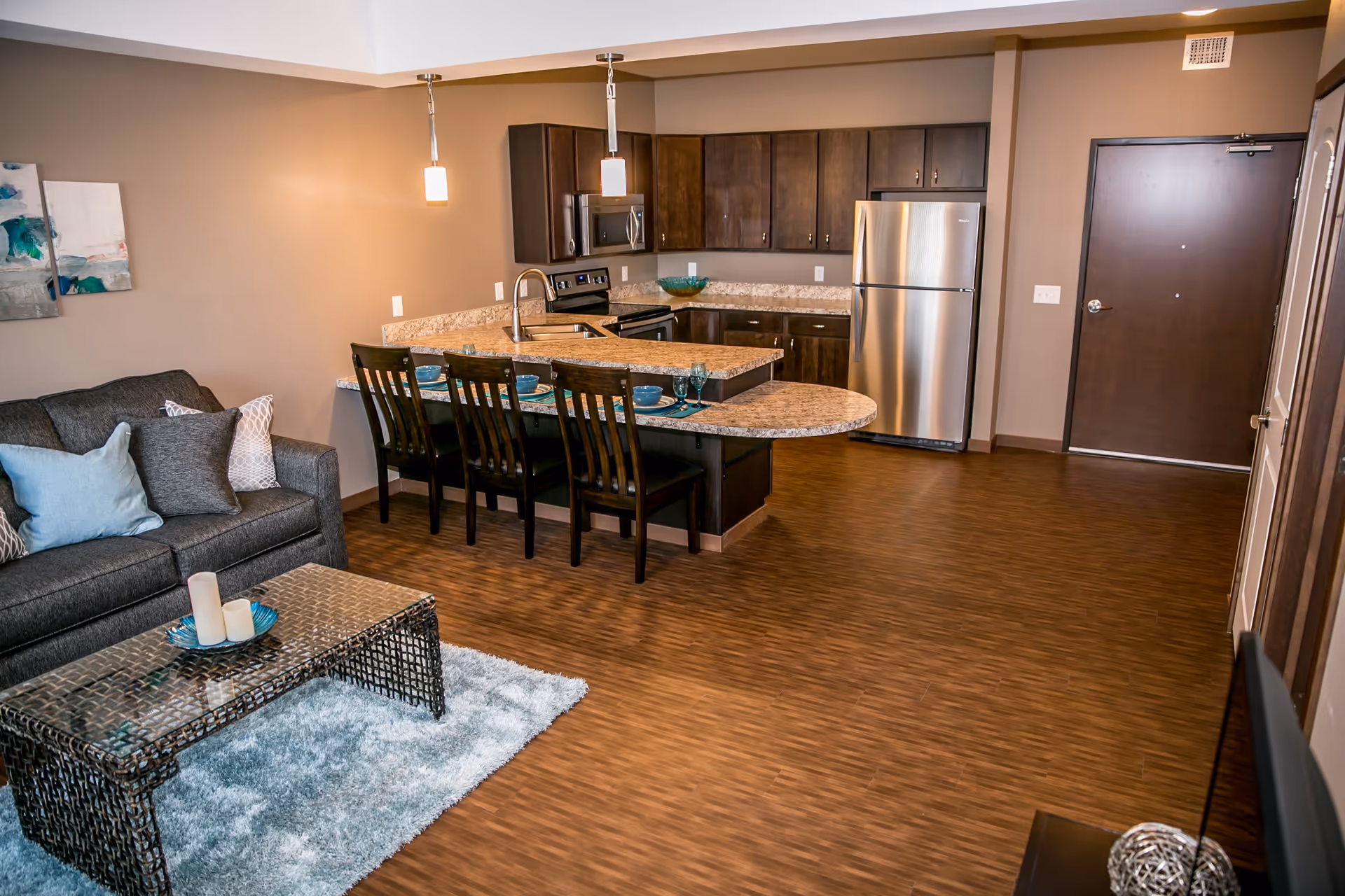 Interior view of a modern living space featuring a kitchen with dark wood cabinets, stainless steel refrigerator, stove, and microwave. A granite countertop island with four wooden chairs is set with blue dishes and glasses. Adjacent to the kitchen is a living area with a gray sofa adorned with various pillows, a glass coffee table on a light blue rug, and wall art. The room has wood flooring and beige walls.