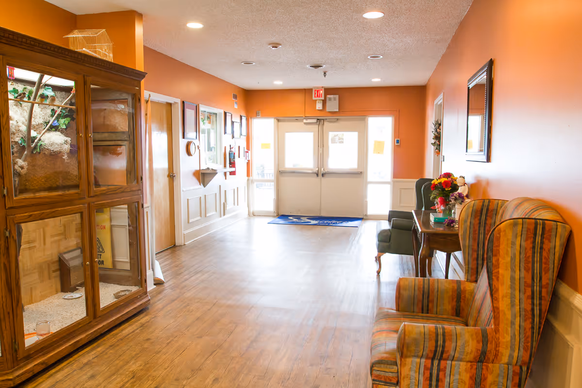 A well-lit hallway with orange walls and wooden flooring. On the right side, there are two upholstered armchairs, a small wooden table with a flower arrangement, and a mirror on the wall. On the left side, there is a large wooden cabinet with glass doors containing a birdcage. At the end of the hallway, there are double glass doors with an exit sign above them.