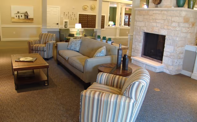 A cozy living room area in a senior living facility featuring a beige sofa with two patterned pillows, two striped armchairs, a wooden coffee table with a decorative plate, a side table with decorative items, and a stone fireplace. The background shows a reception desk and wall decor.