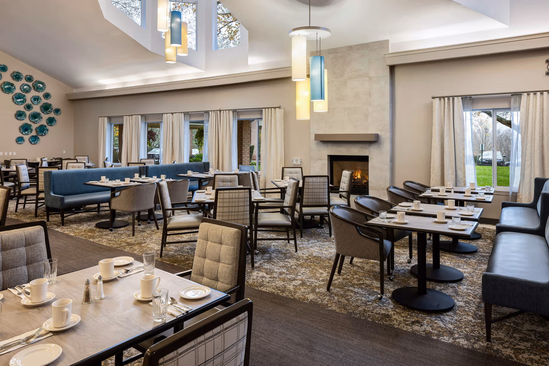 A spacious dining room in a senior living community with multiple tables set with white cups, plates, and silverware. The room features comfortable chairs and blue cushioned bench seating. Large windows with curtains allow natural light to enter, and a modern fireplace is visible on one wall. Decorative blue plates are arranged on another wall, and pendant lights hang from the ceiling.
