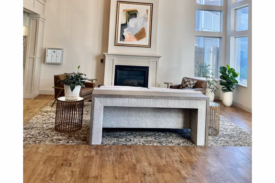 A bright and spacious living room area with a large window letting in natural light. The room features a fireplace with abstract art above it, a light-colored sofa facing away, two brown armchairs, a patterned rug, and several potted plants. The floor is wooden, and the walls are painted white.