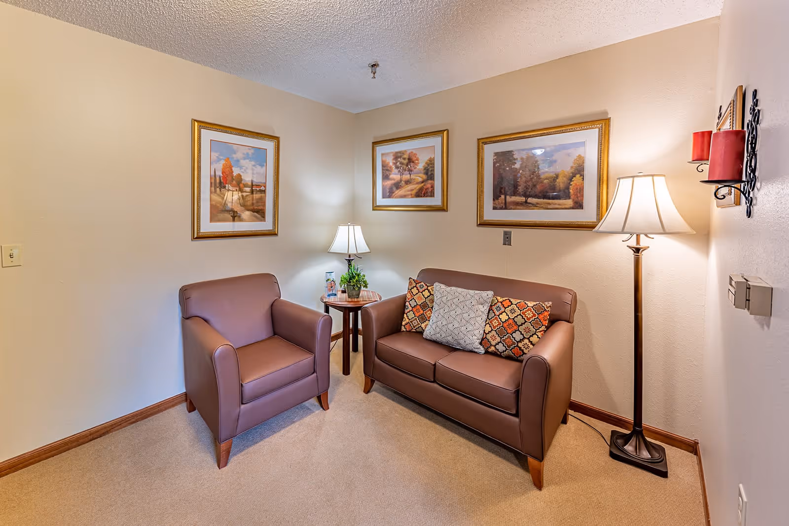 A cozy sitting area in a senior living facility featuring a brown leather armchair and a matching two-seater sofa with decorative pillows. Between them is a small round wooden side table with a lamp and a small plant. The walls are adorned with three framed landscape paintings, and a floor lamp with a white shade stands next to the sofa. The room has beige walls and carpeted flooring.
