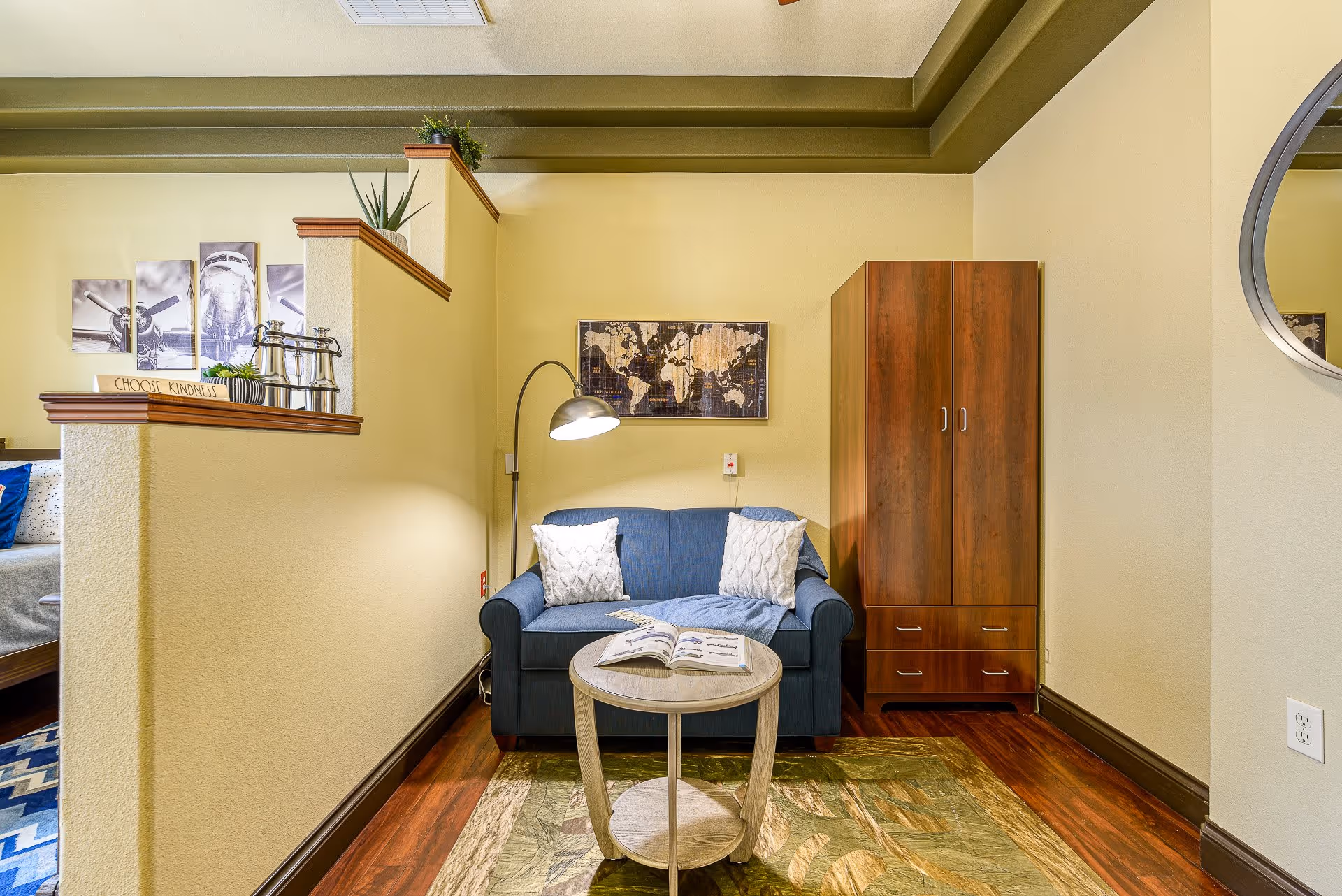 A cozy living area in a senior living facility featuring a blue loveseat with two white pillows and a blue throw blanket. In front of the loveseat is a round wooden coffee table with an open book on it. To the right of the loveseat is a wooden wardrobe. The walls are painted light yellow with a green ceiling border. A floor lamp with a curved neck is positioned behind the loveseat, and a world map artwork hangs on the wall above it. On the left side, a half wall with decorative plants and a sign that says 'CHOOSE KINDNESS' partially separates this space from a bedroom area.