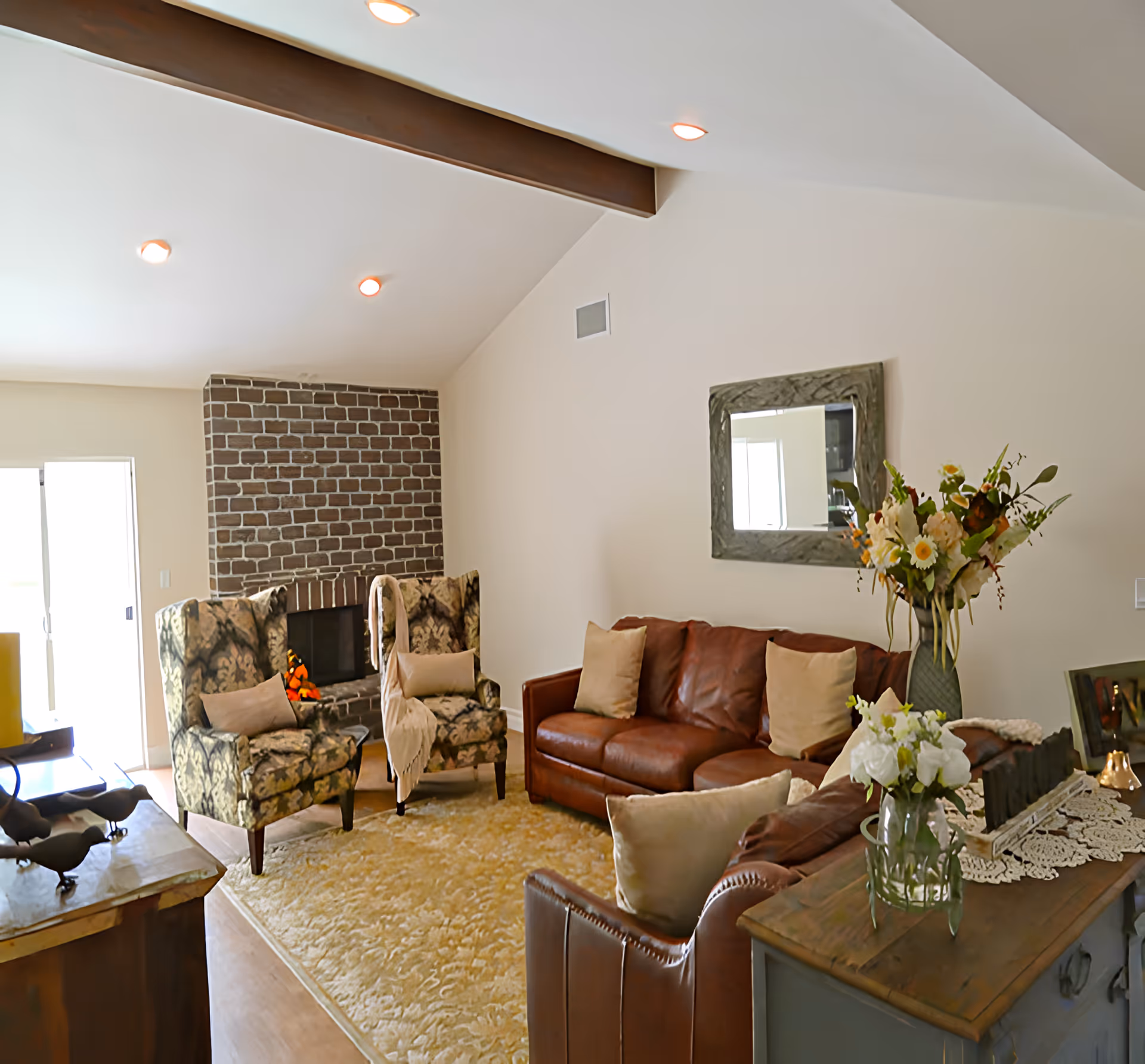 A cozy living room with a brown leather sofa and two patterned armchairs arranged around a brick fireplace. The room features a beige area rug, a wooden sideboard with vases of flowers, a decorative mirror on the wall, and recessed ceiling lights.