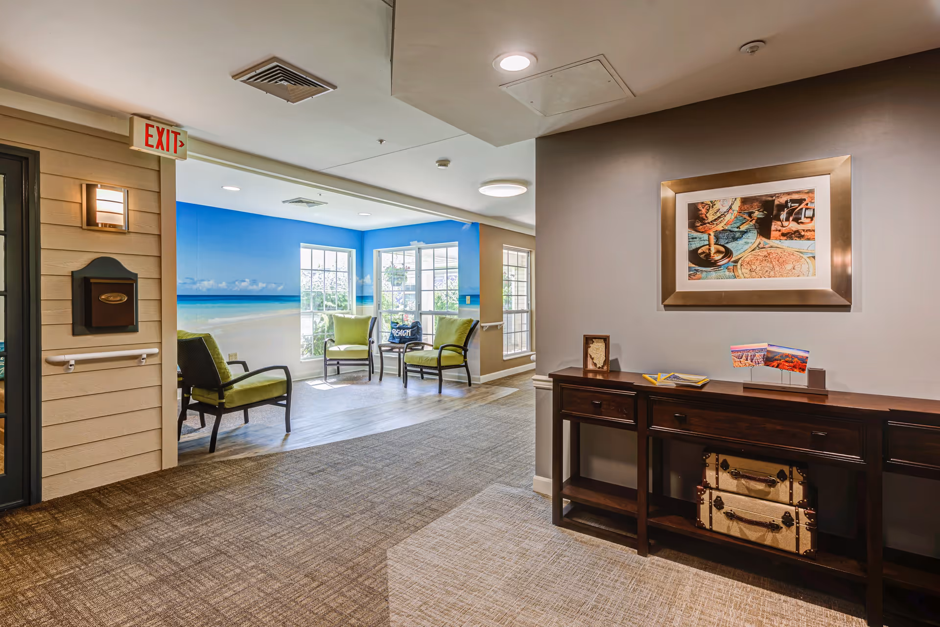 Interior view of a senior living facility hallway with a seating area featuring green cushioned chairs near large windows. The walls have a beach-themed mural, and there is a dark wooden console table with decorative items and framed pictures on the right side. An exit sign is visible above a door on the left.