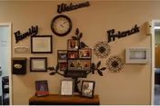 Interior wall decorated with a clock, framed certificates, family photos arranged in a tree-shaped frame, and decorative words 'Family', 'Welcome', and 'Friends'. Below the wall decorations is a wooden table with additional framed photos and documents.