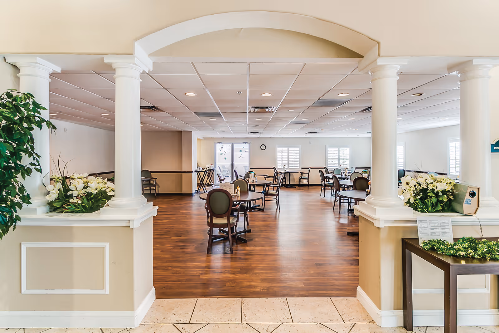 A spacious dining room in Cedar Creek Assisted Living with wooden floors, multiple round tables and chairs, white columns framing the entrance, and large windows letting in natural light. There are decorative plants and flowers on either side of the entrance.