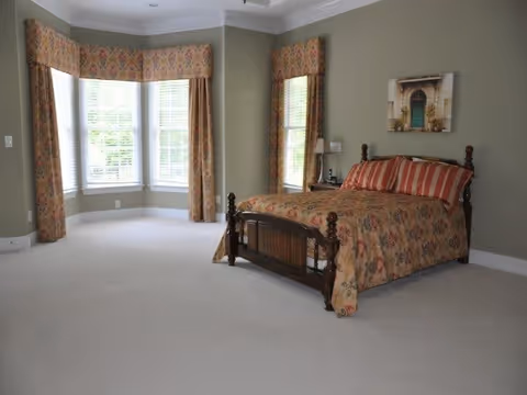 A bedroom with a wooden bed frame and floral patterned bedspread and pillows. The room has light green walls, a large bay window with floral curtains, and a framed picture above the bed. There is a small bedside table with a lamp next to the bed.