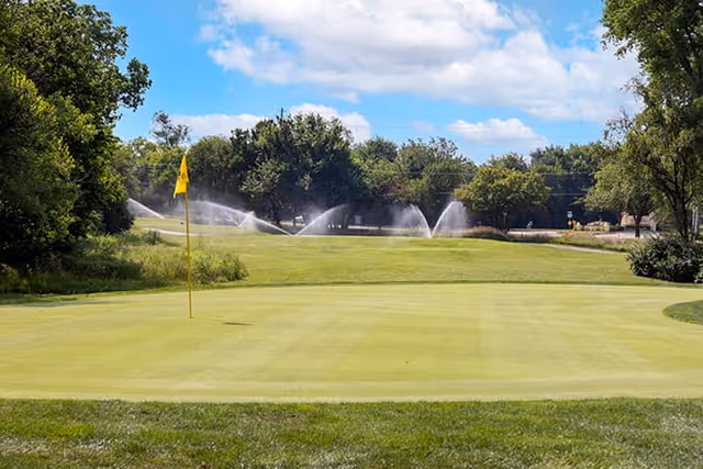 A golf green with a yellow flag and sprinklers watering the fairway against a backdrop of trees and a partly cloudy sky.