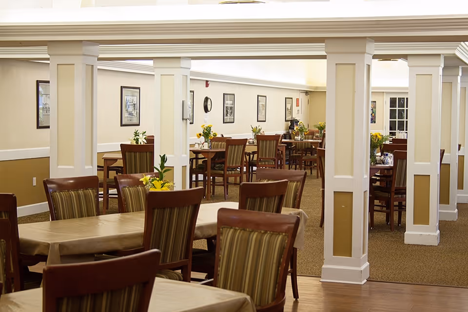 Interior view of a senior living facility dining room with multiple wooden tables and chairs arranged neatly. The room features beige walls with white trim, decorative columns, framed pictures on the walls, and small flower arrangements on the tables.