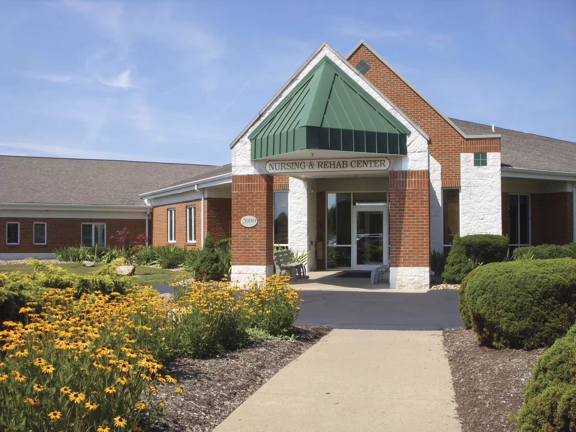 Exterior view of a nursing and rehab center building with a green peaked roof over the entrance. The building is made of red brick and white stone. There is a paved walkway leading to the entrance, surrounded by well-maintained landscaping with yellow flowers and green bushes. The sky is clear and blue.
