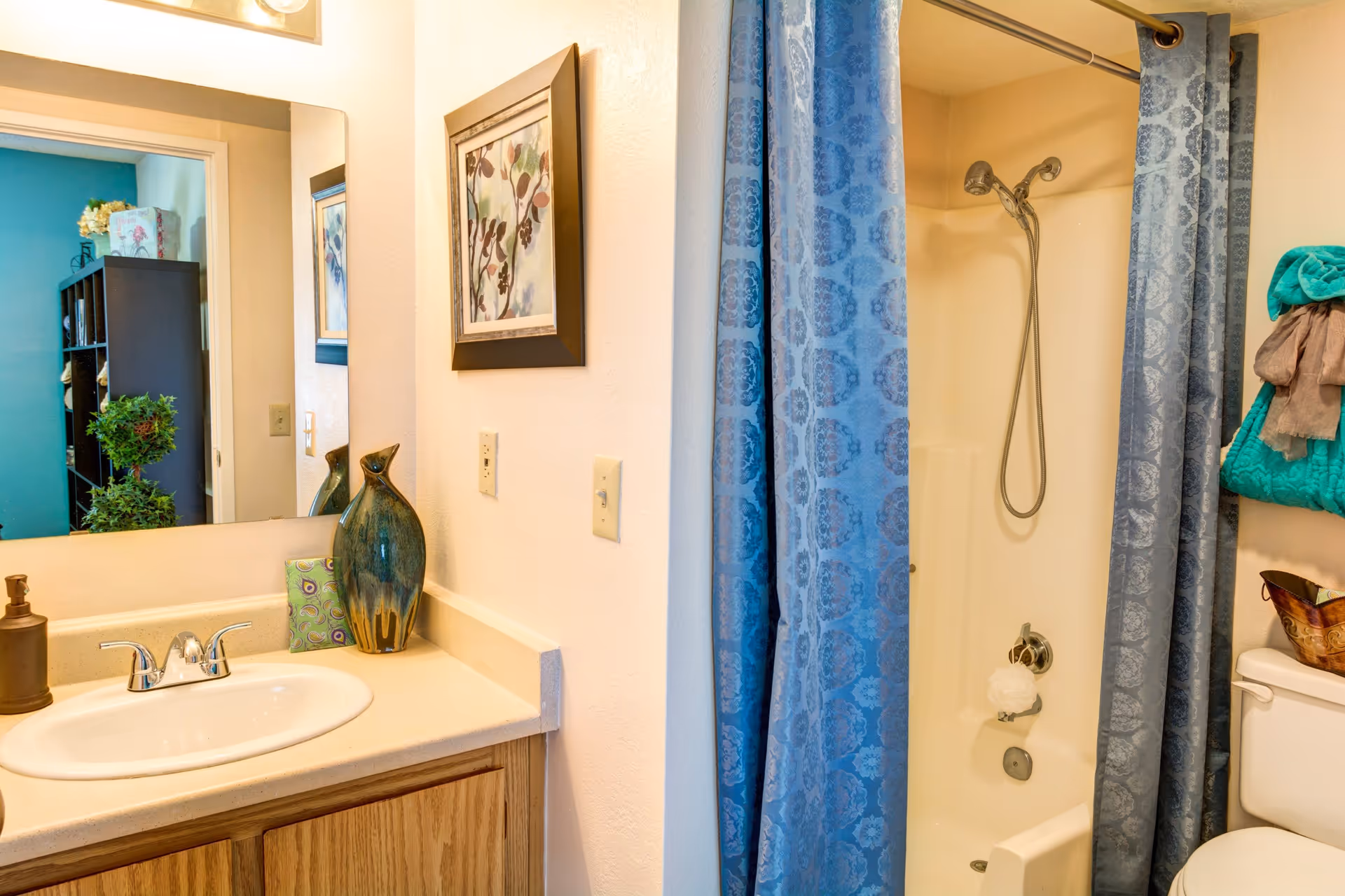 A bathroom with a sink and countertop on the left, a large mirror above the sink, a decorative vase and soap dispenser on the counter, a framed picture on the wall, and a shower with a blue patterned curtain on the right. A toilet with folded towels is partially visible next to the shower.