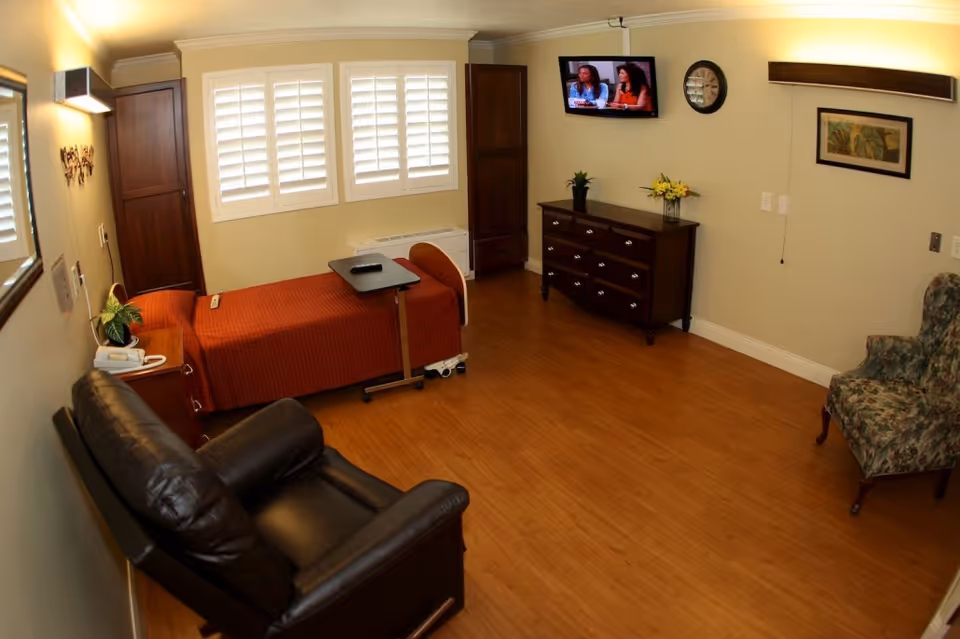 A cozy room with a single bed covered in an orange blanket, a small overbed table, a dark leather recliner, a floral upholstered armchair, a wooden dresser with a plant and flowers on top, a wall-mounted TV showing two women, a clock, and a framed picture on the wall. The room has wooden flooring and windows with white shutters.