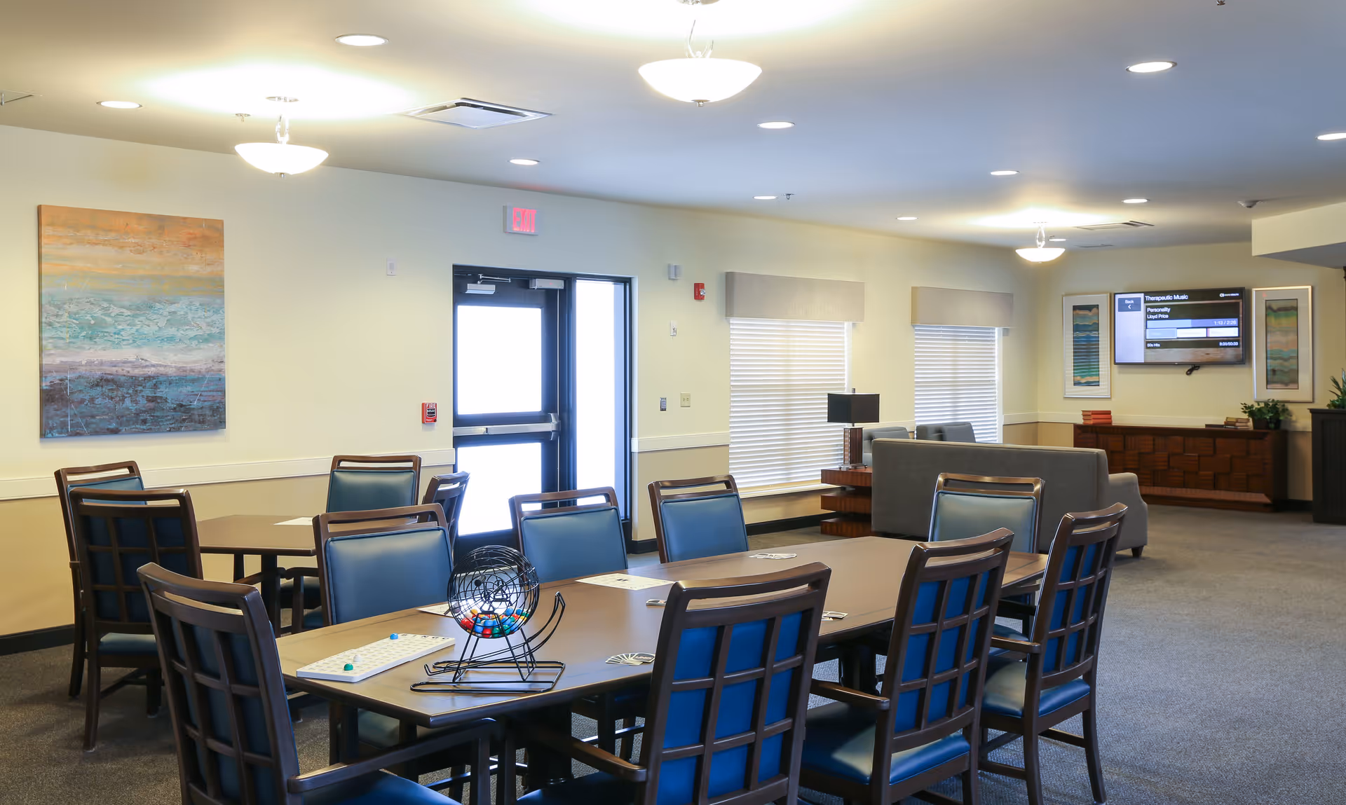 A well-lit common area in Orchard Grove Senior Living featuring a long table with blue cushioned chairs arranged around it. On the table, there is a bingo cage with colorful balls and bingo cards. In the background, there are additional seating areas with sofas, a wall-mounted TV, framed artwork, and large windows with blinds.
