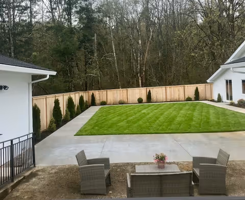 Outdoor courtyard with a rectangular manicured lawn, concrete walkways, patio seating, and a wooden fence backed by trees.